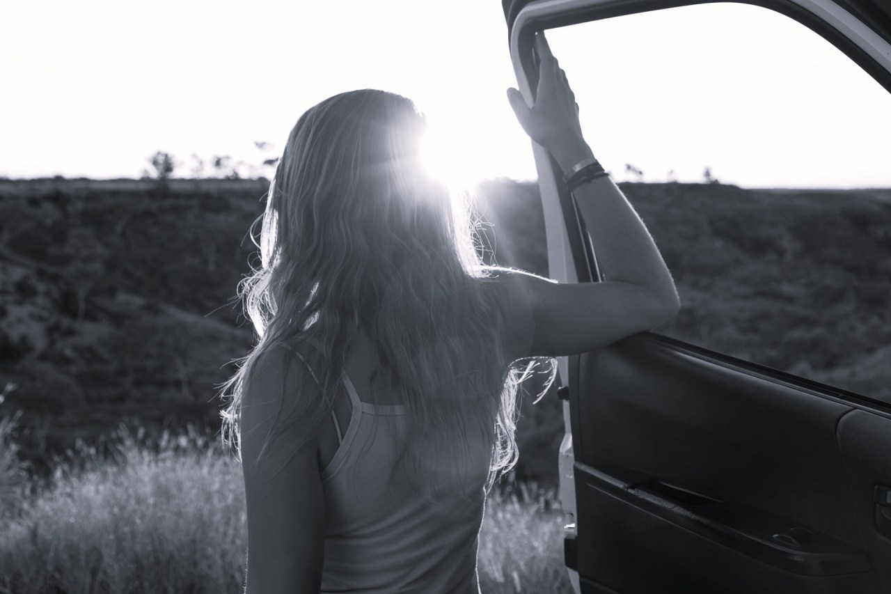 Black and white image of a woman standing beside a car on an open road, representing leaving old ways of thinking behind and moving toward a new life.