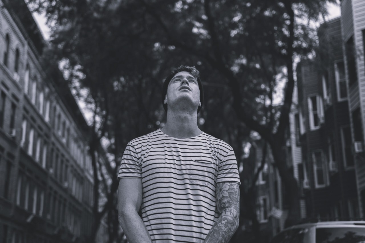 Black-and-white photograph of a man standing alone on a city street, looking upward between tall buildings.