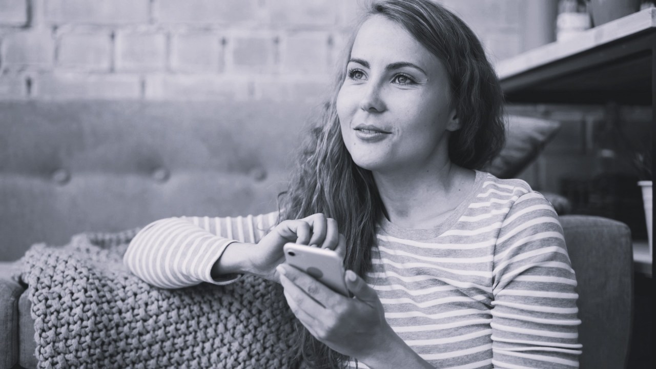 Black-and-white photograph of a woman sitting outdoors at a table, looking thoughtfully into the distance with her hands resting in front of her.
