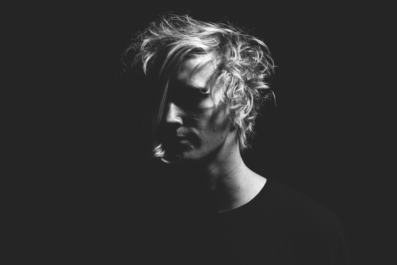 Black-and-white portrait of a person with short hair looking downward, their face partially illuminated against a dark background.